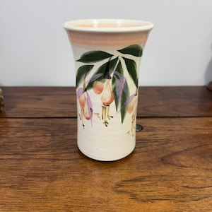 May include: A cream-colored ceramic vase with a flared rim and a soft pink band around the top. The exterior features a hand-painted botanical design of green leaves and drooping pink and purple fuchsia flowers on a smooth, cylindrical body.