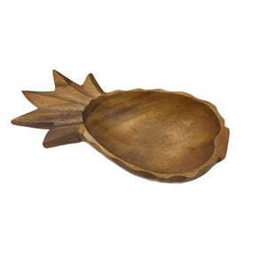 Puede incluir: Un cuenco de madera con forma de piña y acabado natural. El cuenco tiene un diseño tallado y es ideal para servir aperitivos o como pieza decorativa. Las dimensiones del cuenco son de aproximadamente 25 cm de largo y 15 cm de ancho.