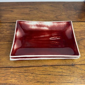 Puede incluir: Un plato rectangular de cerámica con un esmalte rojo brillante profundo y un borde blanco. El centro del plato presenta un diseño en relieve de tres tallos de trigo. El plato tiene una superficie lisa y reflectante con bordes ligeramente elevados.