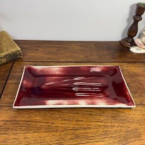 May include: A rectangular ceramic tray featuring a glossy, deep red glaze with a white border. The center of the tray displays a subtle, embossed fork design, creating a textured surface within the smooth, reflective finish of the piece.