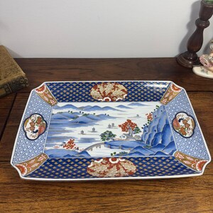May include: A rectangular ceramic serving platter featuring a blue and white landscape scene with mountains, a bridge, and trees. The border is decorated with a dark blue grid pattern, gold accents, and floral motifs in red and white.