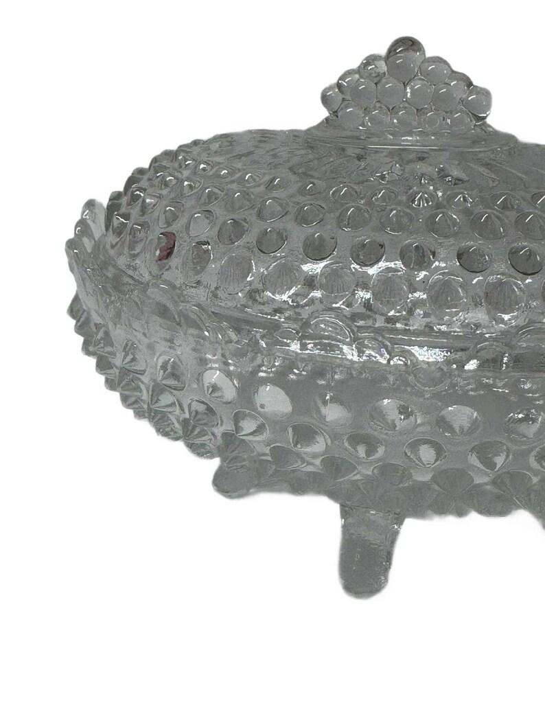 May include: A clear glass candy dish with a lid. The dish has a hobnail pattern and sits on three small feet. The lid has a knob made of stacked glass spheres. The dish is empty and sits against a white background.