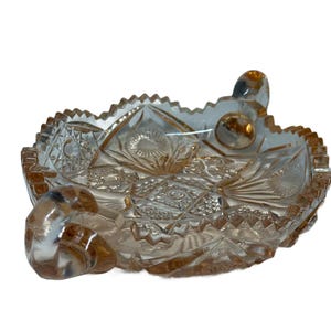 May include: A clear, decorative glass dish with a detailed, cut-glass pattern. The dish has a scalloped edge and two rounded handles. The glass has a slight amber tint, and the design includes starburst and geometric shapes.