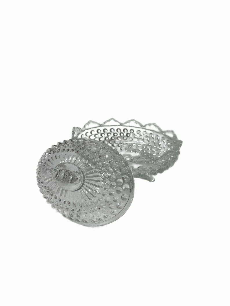 May include: A clear, vintage glass candy dish with a lid. The dish and lid are decorated with a textured pattern of small, raised dots and a scalloped edge. The lid is partially open, revealing the interior of the dish.