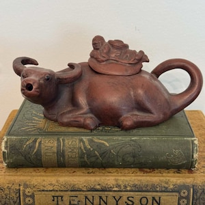 以下が含まれることがあります： 水牛の形をした茶色のセラミックティーポット。背中には人物がいます。ティーポットは、金色の文字が入ったアンティーク本の山の上に置かれています。ティーポットのハンドルは湾曲したループです。