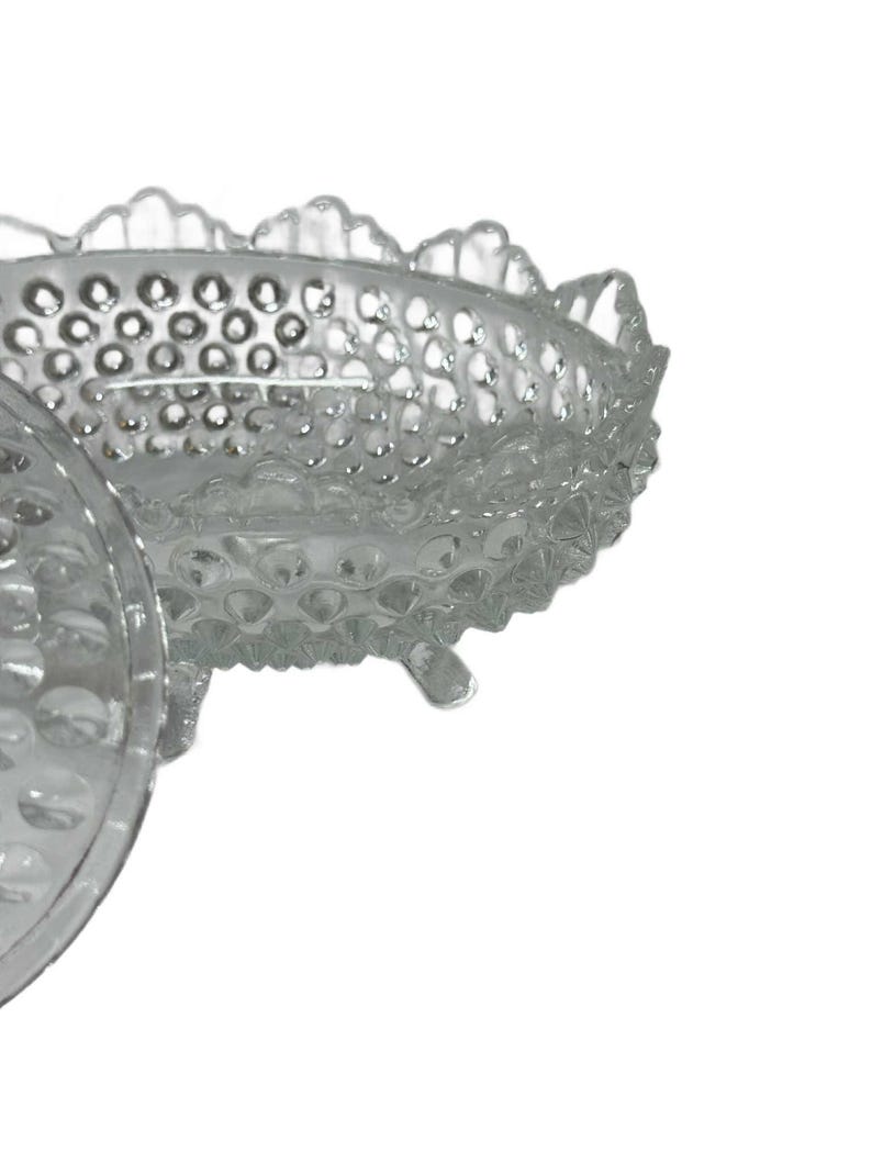 May include: Clear glass bowl with a hobnail pattern and scalloped rim. The bowl has three small feet and a lid. The glass is transparent, allowing light to pass through. This vintage bowl is suitable for serving or display.