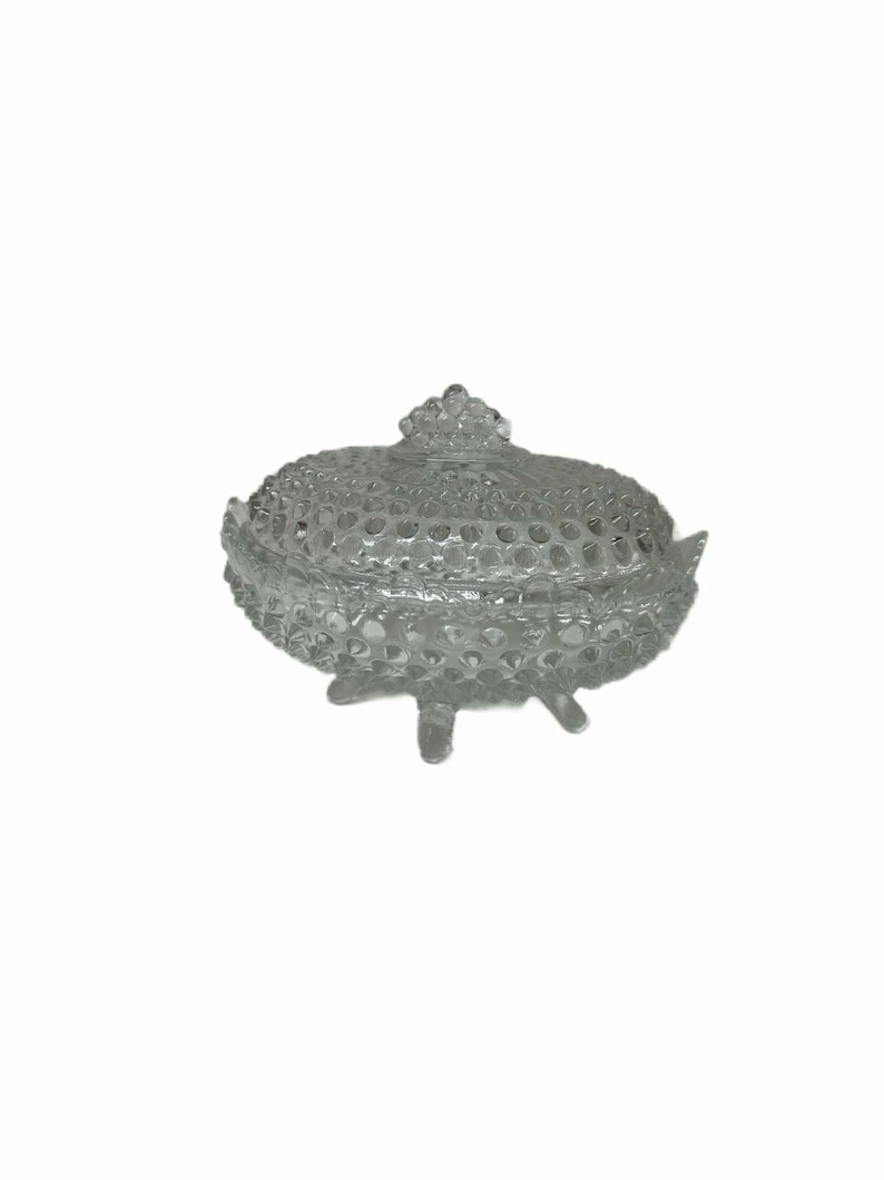 May include: A clear, glass candy dish with a lid. The dish has a textured, hobnail pattern and sits on three small feet. The lid is topped with a decorative finial. The dish is designed to hold small candies or other treats.