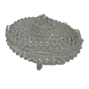 May include: Clear glass oval-shaped covered dish with a textured, bubble-like pattern. The lid features a cluster of small glass beads as a handle. The dish has a scalloped edge and rests on three small feet. A decorative piece for the home.