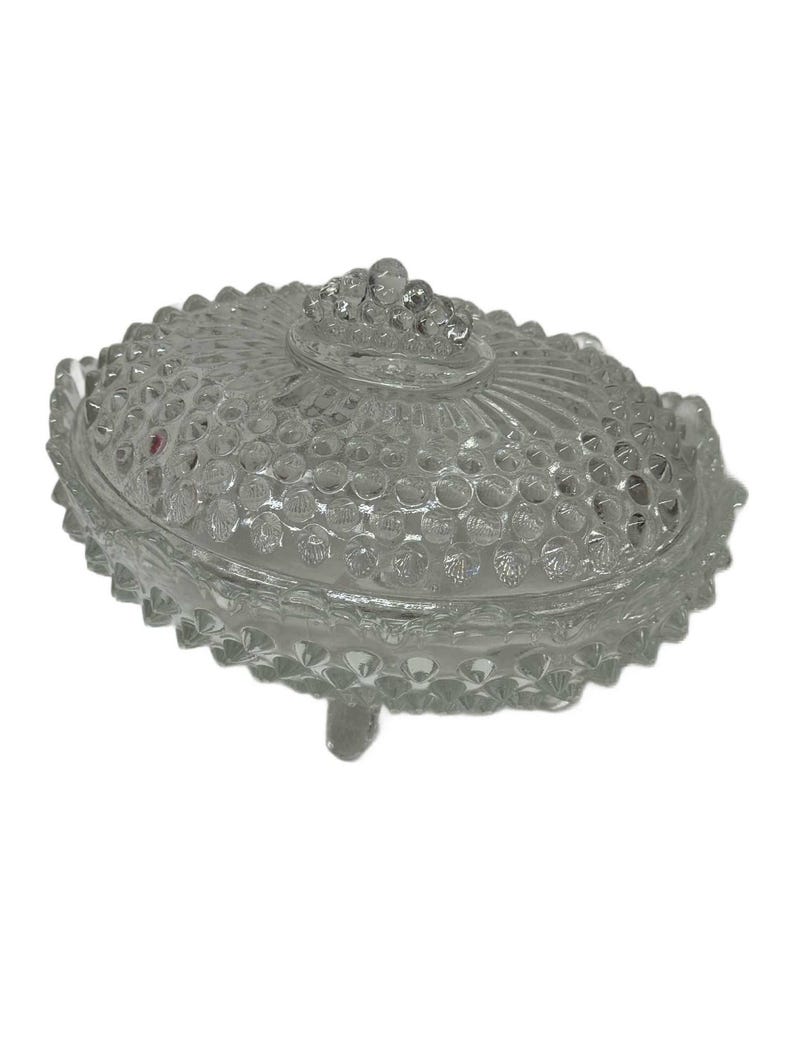 May include: Clear glass oval-shaped covered dish with a textured, bubble-like pattern. The lid features a cluster of small glass beads as a handle. The dish has a scalloped edge and rests on three small feet. A decorative piece for the home.