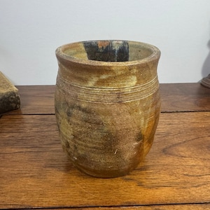 May include: A cylindrical ceramic vessel with a mottled brown and tan glaze. The exterior features horizontal incised lines near the rim, with a dark, charred-looking patch near the top edge and a textured, earthy finish throughout the body.