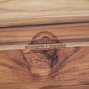 May include: A round wooden serving board with a natural wood finish. The board has a light brown color and a darker brown center. The board is engraved with the text "Wilderness & Treasures".