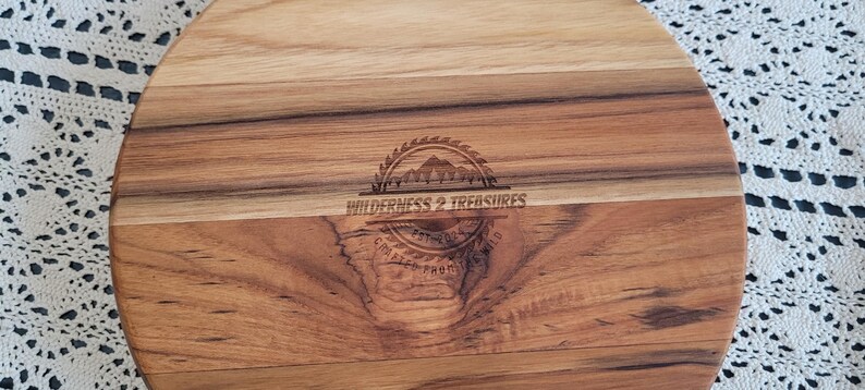 May include: A round wooden serving board with a natural wood finish. The board has a light brown color and a darker brown center. The board is engraved with the text "Wilderness & Treasures".