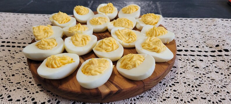 May include: A wooden platter filled with deviled eggs. The eggs are white with a yellow yolk filling. The platter is on a white crocheted tablecloth.