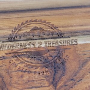 May include: A wooden cutting board with a logo that reads "By Wilderness 2 Treasures Est. 2021 Crafted From The Wild".