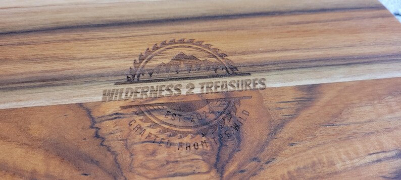 May include: A wooden cutting board with a logo that reads "By Wilderness 2 Treasures Est. 2021 Crafted From The Wild".