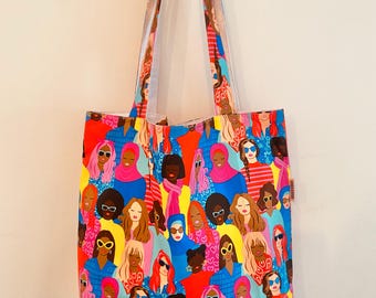 Può includere: Una borsa tote colorata con un motivo di donne che indossano occhiali da sole e vari copricapi. La borsa ha lunghi lacci e è realizzata in un tessuto resistente.