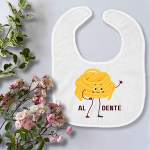 May include: White baby bib with a cartoon illustration of a smiling bundle of pasta with arms and legs. The pasta is yellow and has a red mouth. The text "AL DENTE" is printed below the pasta.