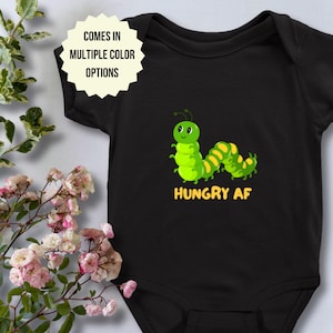 May include: Black baby bodysuit with a cartoon caterpillar design. The green caterpillar has yellow accents and the text "HUNGRY AF" in yellow. The bodysuit also has the text "COMES IN MULTIPLE COLOR OPTIONS" in a white starburst.