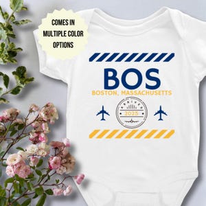 May include: A white baby grow with a blue and yellow airport arrival stamp design. The stamp says "Arrived 2025" and has a small aeroplane icon. The grow also has the text "BOS Boston, Massachusetts" and two aeroplane icons.