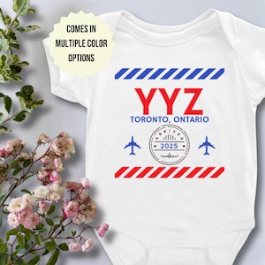 Puede incluir: Mono blanco para bebé con un diseño de llegada al aeropuerto en rojo, blanco y azul. El diseño incluye el texto "YYZ TORONTO, ONTARIO" y un sello que dice "ARRIVED 2025".