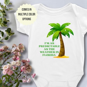 May include: White baby bodysuit with a green and brown palm tree graphic and the text "I'M AS PREDICTABLE AS THE WEATHER IN FLORIDA." The text "COMES IN MULTIPLE COLOR OPTIONS" is also displayed.