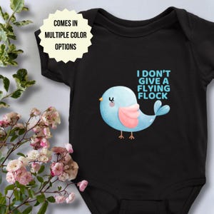 May include: Black baby bodysuit with a blue bird illustration and the phrase "I DON'T GIVE A FLYING FLOCK." The bird has pink wings and a yellow beak. The text "COMES IN MULTIPLE COLOR OPTIONS" is also displayed.