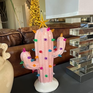 May include: A pink ceramic cactus figurine with colorful lights and a gold star topper. The cactus has a white base and is decorated with various colored lights. The figurine is a decorative item.