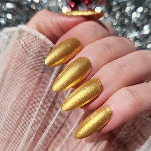 May include: Close-up of a hand with long, gold-coloured fingernails. The nails have a shiny, metallic finish. The background is blurred, with hints of silver and red.