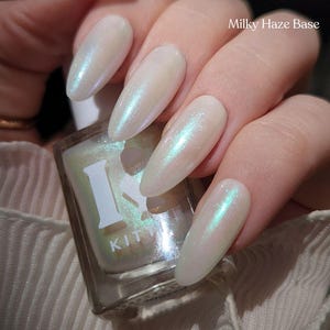 May include: Close-up of long, oval-shaped nails painted with a pearlescent, milky white nail polish. The polish has a subtle iridescent shimmer, reflecting hints of blue and green. A bottle of nail polish with the text "Milky Haze Base" is visible.