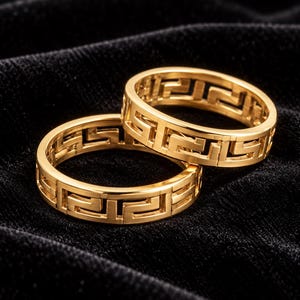 Può includere: Due anelli color oro caratterizzati da un motivo a chiave greca traforato continuo lungo l'intera fascia. Gli anelli presentano una finitura metallica lucida e una larghezza uniforme, con un complesso design geometrico che si ripete.
