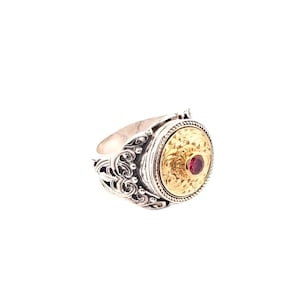 May include: A silver ring with a gold-coloured circular centrepiece. The gold centrepiece features an ornate design and a small, round, red gemstone. The ring has a detailed, swirling pattern on the band.