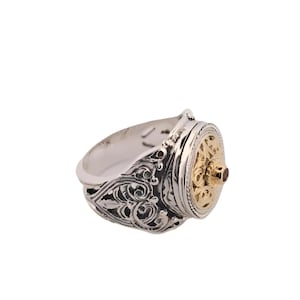 Puede incluir: Un anillo de plata con detalles de filigrana ornamentados en la banda. El anillo presenta una pieza central circular de color dorado con una pequeña gema roja. El diseño es intrincado y detallado, de estilo vintage o antiguo.