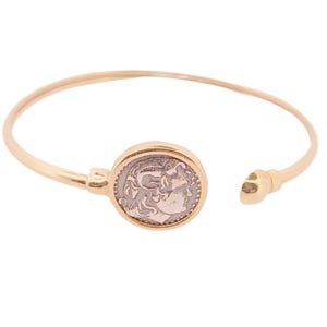 Puede incluir: Brazalete de puño abierto en tono dorado con un medallón circular con un diseño de perfil detallado. El brazalete tiene un acabado liso y pulido y un estilo clásico. El diseño abierto facilita su uso.