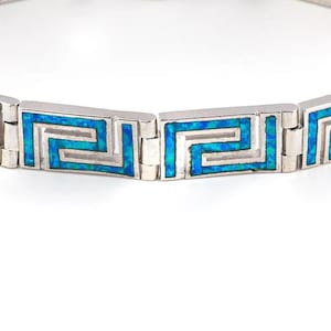 May include: A silver-toned bracelet featuring a Greek key design. The bracelet has rectangular links with a blue and silver pattern. The links are connected by smaller, silver-toned rectangular pieces. The bracelet is displayed against a white background.