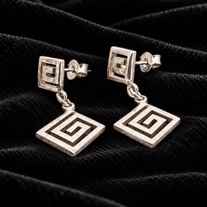May include: A pair of silver-toned drop earrings featuring a square stud top and a larger square dangling pendant, both decorated with a cut-out Greek key pattern. The earrings include standard post-back closures with metal butterfly-style backs.