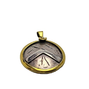 Può includere: Un ciondolo rotondo con bordo e anello dorati. Il centro del ciondolo presenta un disegno argentato di uno scudo. Il disegno dello scudo ha un bordo dorato. Il ciondolo è probabilmente in metallo.