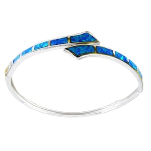 Può includere: Un bracciale a cerchio argentato con intarsi di opale blu. Il bracciale presenta un design unico con una forma curva e aperta. Le pietre di opale blu sono disposte in un motivo rettangolare, creando un contrasto con l'argento.