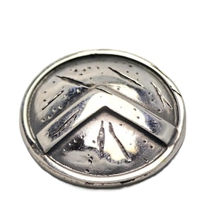 Könnte beinhalten: Ein silberfarbener, runder Schild mit einem markanten Chevron-Design. Der Schild hat ein verwittertes Aussehen mit geätzten Linien und kleinen Punktdetails, was auf eine antike oder historische Ästhetik hindeutet. Der Schild ist wahrscheinlich ein Dekorationsartikel oder ein Schmuckstück.