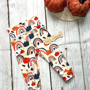 May include: Cream-colored leggings with a fall-themed pattern of rainbows, pumpkins, and leaves. The leggings are displayed on a white wooden surface, with decorative pumpkins in the background. The text "@Hi Donna!" is visible on a wooden tag.