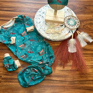 May include: A teal blue knotted baby gown with a dragon and ship pattern. The gown is paired with a matching teal blue knotted hat and a pair of teal blue knotted mittens. The gown is laid out on a wooden surface with a dreamcatcher and feathers.