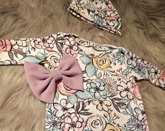 Puede incluir: Un conjunto de ropa para bebé con un mono de manga larga con estampado floral y un gorro a juego con estampado floral. El mono tiene un lazo lavanda en la parte delantera.