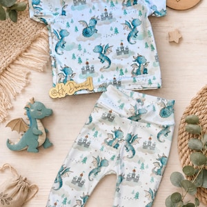 Könnte beinhalten: Ein Set aus einem kurzärmeligen T-Shirt und einer Hose mit einem sich wiederholenden Muster aus blauen Drachen, grauen Burgen und grünen Tannenbäumen auf weißem Grund. Das Set besteht aus einem Oberteil mit Rundhalsausschnitt und einer Hose.
