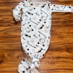 May include: A white baby gown and matching hat set. The gown features a golf-themed pattern with golf carts, clubs, and tees in black and gray. The hat is a knotted style. The set is designed for newborns.