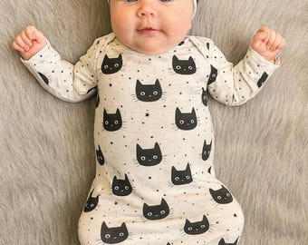 May include: A baby wearing a light-colored onesie and matching headband, both patterned with black cat faces and small stars. The onesie is long-sleeved and knotted at the bottom. The baby is lying on a gray, furry surface.