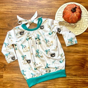 May include: A white long-sleeved baby outfit with a teal trim and a matching headband. The outfit is patterned with cartoon ghosts wearing witch hats and holding cauldrons. The word "BOW" is printed on the outfit.