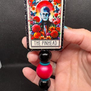 May include: A retractable badge reel featuring a rectangular design with the text "THE PINHEAD". The design includes a portrait surrounded by flowers and butterflies. The reel has black, red, and teal beads.