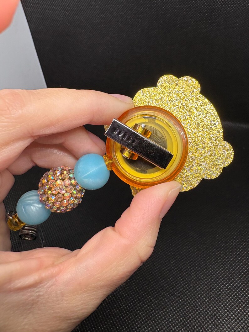May include: A gold glitter charm with a yellow plastic backing and a metal clip. The charm is attached to a beaded necklace with blue and pink beads.