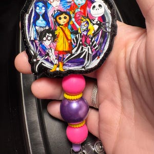 May include: A badge reel featuring a colorful illustration of characters from the animated movie. The badge reel has a black glittery border and is attached to a beaded lanyard with pink, purple, and yellow beads.