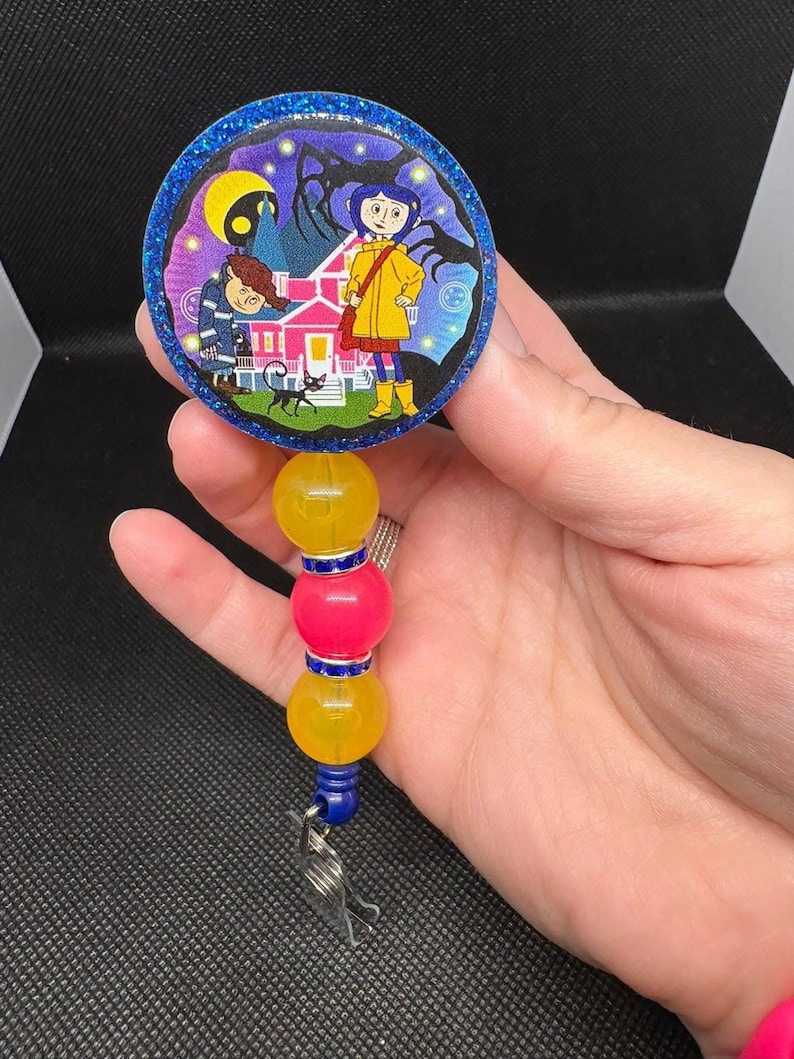 May include: A blue and yellow beaded retractable badge holder with a round, glittery, colorful image of a girl and a boy in front of a pink house. The image is from the movie "Coraline".