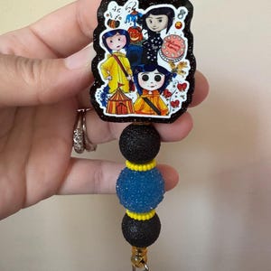 May include: A black and glittery badge reel featuring a colorful illustration of characters from a movie. The badge reel has three decorative beads in black, blue, and yellow. The reel is attached to a clear plastic clip.
